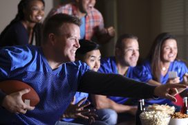 Multi-ethnic group of football fans watching the game together on television at home.  They have snacks, drinks, and sports ball and wear blue team jerseys.  Mid-adult man foreground.