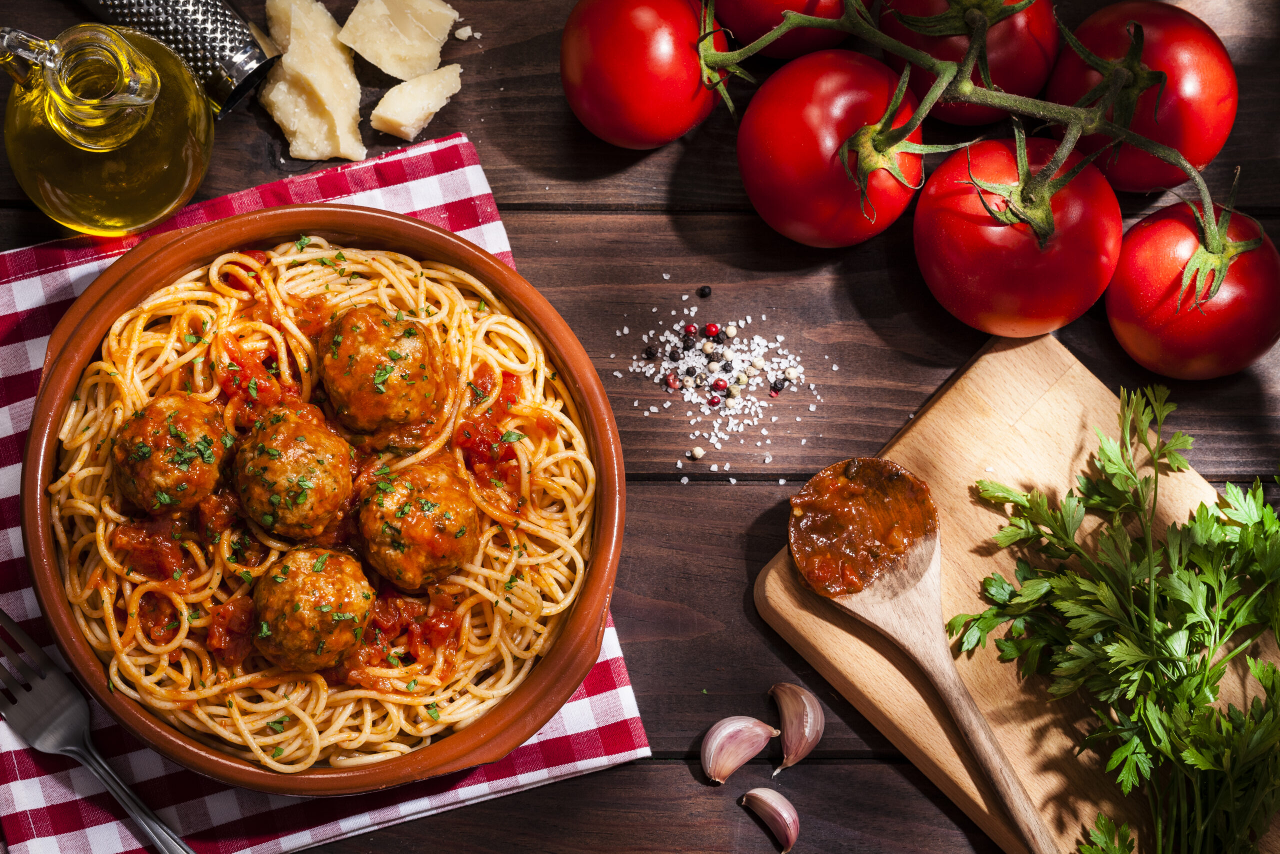 Top view of a plate filled with spaghetti and meatballs with some ingredients for preparation like tomatoes, parsley, garlic, olive oil and Parmesan cheese shot on rustic wooden table. Low key DSRL studio photo taken with Canon EOS 5D Mk II and Canon EF 100mm f/2.8L Macro IS USM